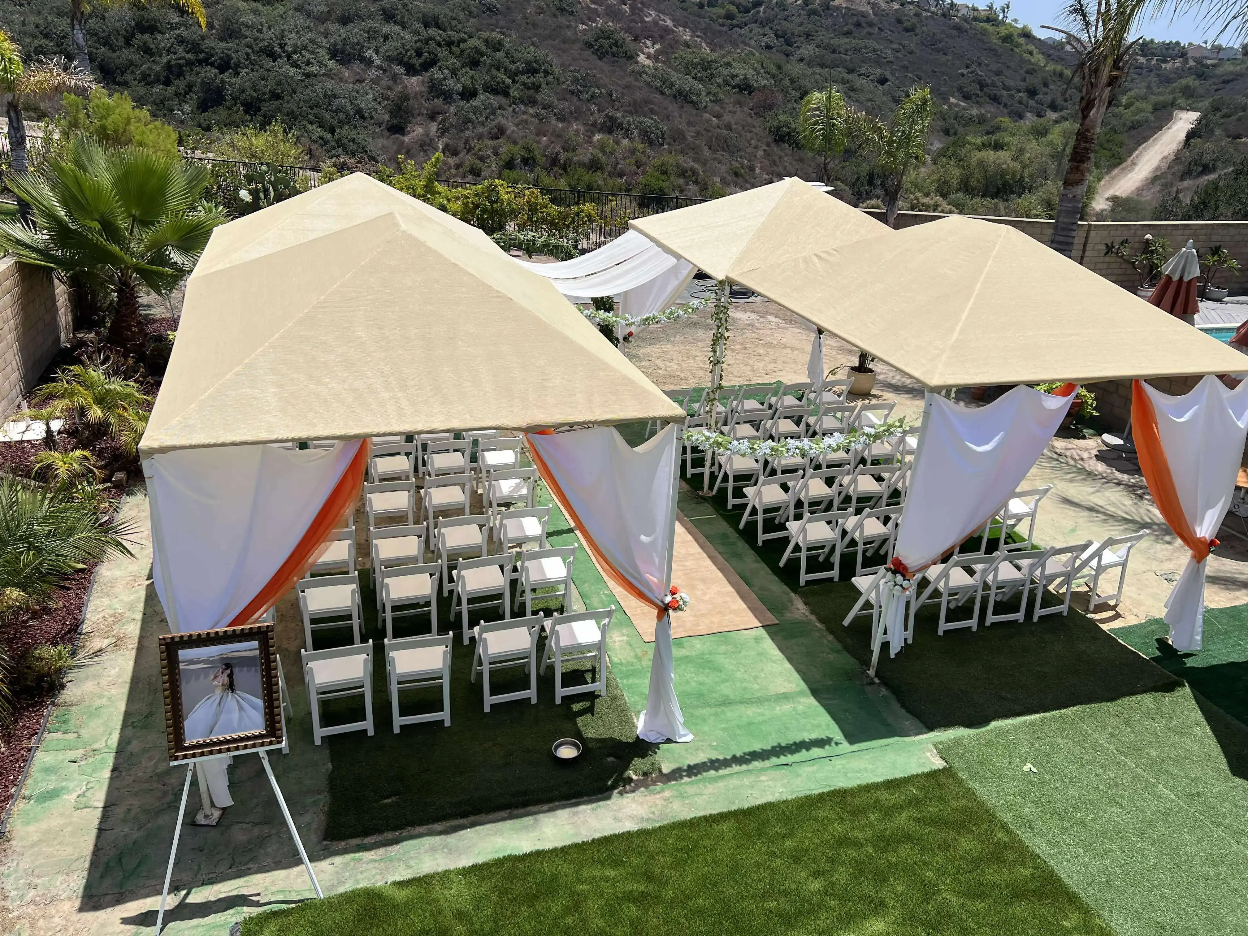 Garden event setup with two beige canopy tents, rows of white chairs, decorative drapes, and a framed picture at the entrance.