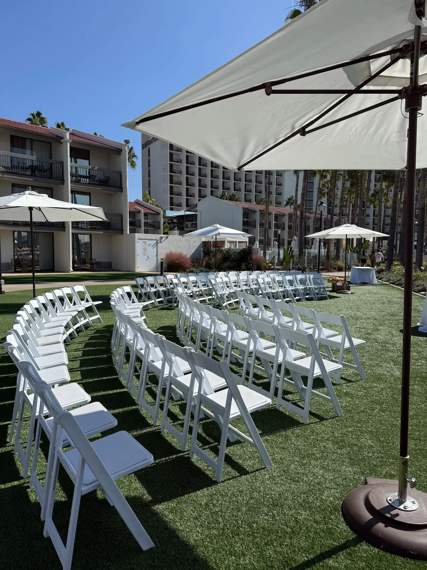 Outdoor event setup with white folding chairs arranged in a semi-circle on grass, large umbrellas, and hotel buildings in the background.