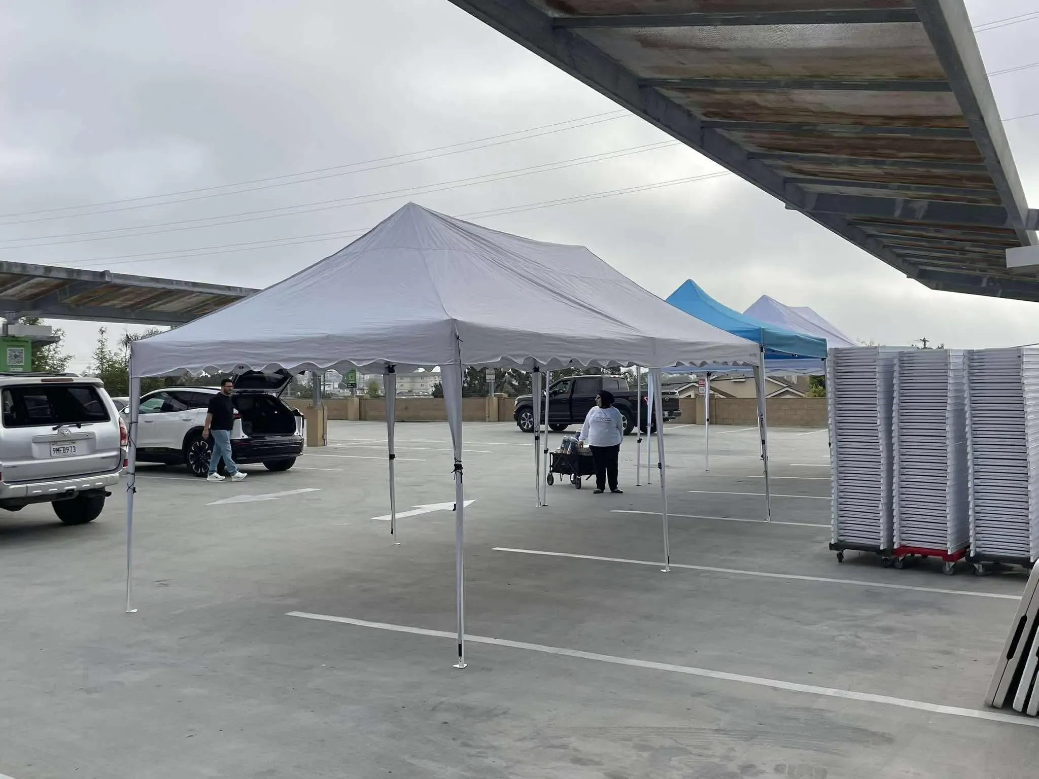 Parking lot setup with white canopy tents, stacked white chairs, and people unloading equipment from vehicles.